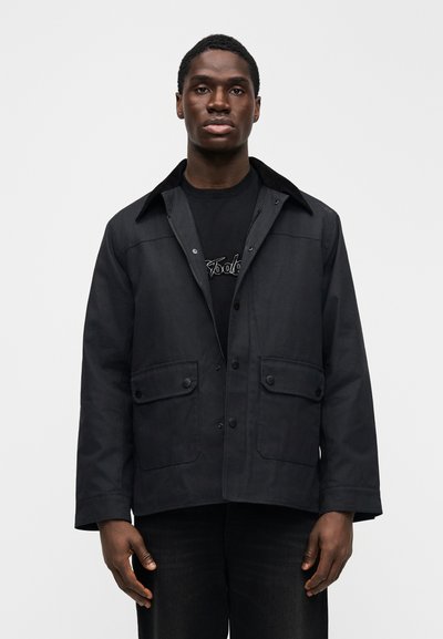 Μαύρο εργασιακό μπουφάν με γιακά, κλείσιμο με κουμπιά μπροστά, δύο τσέπες στο στήθος και λεία υφή υφάσματος. Φοράται πάνω από μαύρο πουκάμισο.