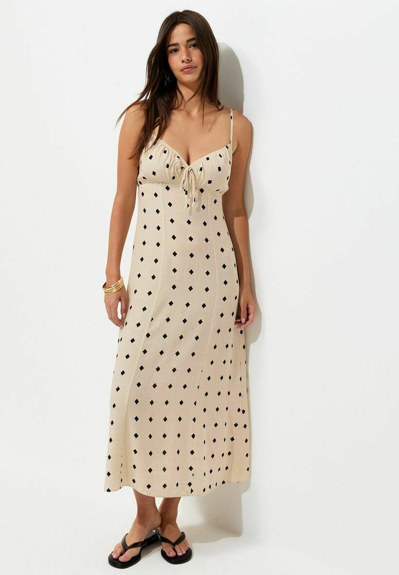 Woman with long dark hair wearing a beige sleeveless midi dress with black diamond patterns and black sandals, standing against a white wall.