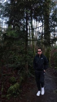 Veste et pantalon imperméables noirs portés avec des baskets blanches, debout sur un sentier de terre entouré d'une forêt dense et de troncs d'arbres moussus.