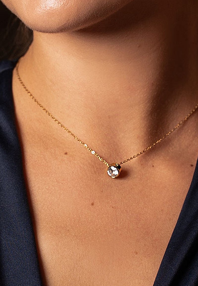 Gold chain necklace with a small round pendant featuring a clear crystal accent, worn against a navy fabric background.