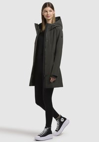 Cappotto impermeabile verde scuro con cappuccio, bottoni e cerniera. Abbinato a leggings neri e sneakers alte nere con suole bianche.
