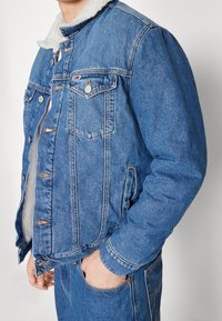 Man med blå denimjacka med vit fleecefoder och matchande blå denimjeans, hand i framfickan mot vit bakgrund.