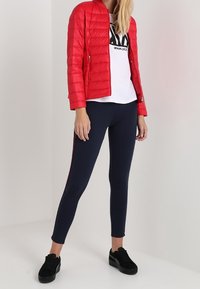 Červená prošívaná bunda, bílý tričko s logem, námořnické legíny s bočními pruhy a černé tenisky. Bunda má zapínání na zip a prošívanou texturu.