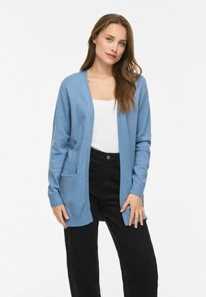 Femme aux longs cheveux bruns portant un cardigan bleu clair ouvert, un haut blanc et un pantalon noir taille haute, debout devant un fond uni.