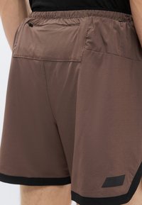 Καφέ αθλητικό σορτς με μαύρη ζώνη μέσης, με ελαστικό επάνω μέρος, πίσω τσέπη και μαύρη λεπτομέρεια στο τελείωμα.