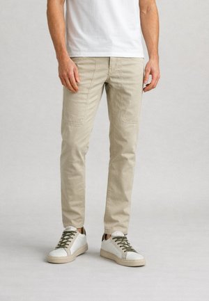 Uomo con pantaloni beige con tasche applicate frontali e sneakers bianche con lacci verdi, in piedi su un pavimento grigio chiaro.