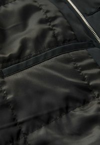 Tejido de chaqueta acolchada negra con un acabado suave y brillante. Presenta un patrón de acolchado diagonal y un bolsillo con un borde suave. Detalle de cremallera plateada.
