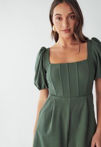 Robe verte à manches bouffantes avec décolleté carré, ornée de plis verticaux et en tissu lisse. Remarquable par sa texture douce et sa coupe structurée.