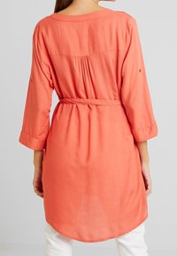 Túnica color coral con mangas largas, cintura ceñida, espalda fruncida y dobladillo redondeado. Confeccionada en una tela ligera y suave.