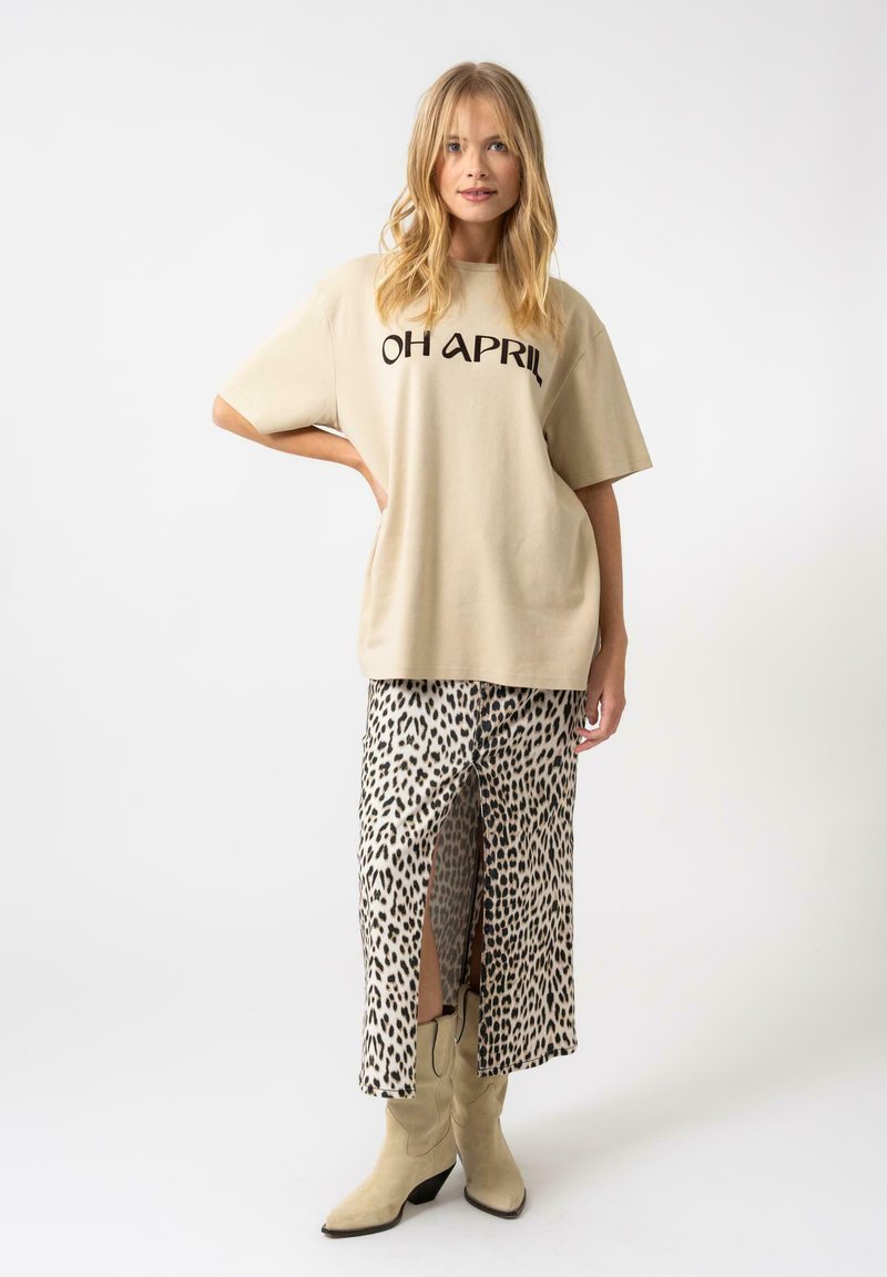 Camiseta oversized beige con el texto "OH APRIL", combinada con una falda de estampado de leopardo con una abertura y botas de cowboy color piel.