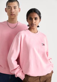 Dos personas llevan sudaderas de gran tamaño en un suave color rosa pastel, con mangas largas y puños. El hombre lleva un collar de cadena; la mujer tiene un parche con un logo.