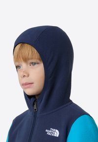 Sötétkék polár cipzáras kapucnis pulóver élénk kék ujjal, elöl cipzárral és logóval a mellrészen. Simogató, puha tapintású anyag.