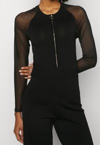 Haut noir à manches longues avec des manches en maille transparente, corps en côte et fermeture éclair sur le devant, arborant un accent métallique circulaire au niveau du col.