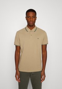 Polo beige in cotone, con colletto classico, dettagli a strisce verdi sul colletto e sulle maniche, e un piccolo logo sul petto.
