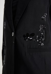 Juodas bleiseris su gėlių sequin siuvinėjimu sidabro ir juodos spalvos. Išskirtinės detalės apima vieną mygtuko užsegimą ir šonines kišenes.