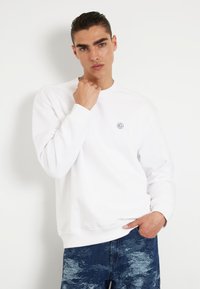 Valge dressipluus ümmarguse kaelusega ja pikkade varrukatega, millel on rinna peal väike logo. Seljas sinised mustrilised teksad.