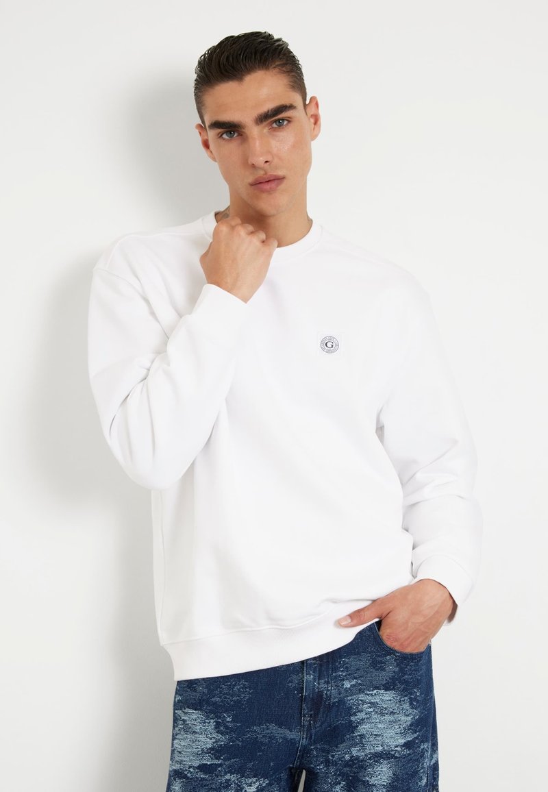 Valge dressipluus ümmarguse kaelusega ja pikkade varrukatega, millel on rinna peal väike logo. Seljas sinised mustrilised teksad.