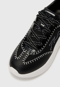 Baskets noires avec une tige en tissu texturé, des coutures blanches contrastantes et un accent en plastique noir lisse au niveau des lacets. Semelle blanche.
