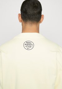 Light yellow sweatshirt featuring a circular logo on the back with "Nike Athletic Club 1972" text in dark blue. Soft texture and casual design.