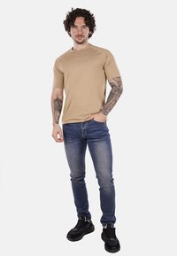 T-shirt beige a maniche corte con collo tondo, abbinato a jeans slim fit blu e scarpe da ginnastica nere. Il modello sta in piedi con le mani giunte.