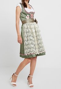 Robe verte avec un tablier en dentelle, des accents floraux et une ceinture en satin. Comprend un corsage ajusté, un col rond et un ourlet au niveau du genou. Chaussures blanches.