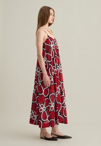Rotes Kleid mit Blumenmuster und weißen Akzenten, ärmelloses Design, leichter Stoff und fließende Silhouette. Kombiniert mit schwarzen Ballerinas.
