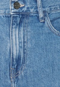 Pohled zepředu na modré džíny z denimu zobrazující knoflík, pas, poutko na opasek, kapsu a detailní švy.