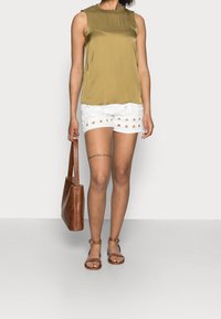 Top sin mangas de color oliva combinado con pantalones cortos blancos texturizados que presentan recortes. Un bolso de hombro de cuero marrón y sandalias marrones con tiras completan el look.