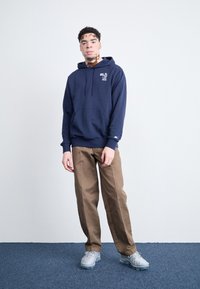 Sweat-shirt bleu marine avec poche avant et détail de logo, associé à un pantalon marron clair. Le modèle se tient sur un tapis bleu avec un arrière-plan neutre.