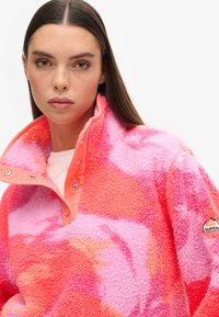 Fleece-sweater i levende pink og orange nuancer med høj krave, trykknapper og et mærke på ærmet. Blød tekstur.