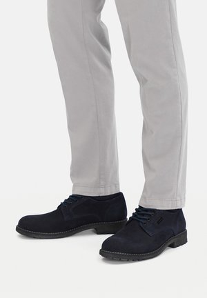 Granatowe zamszowe buty o zaokrąglonych czubkach, czarne sznurowadła i kontrastujące przeszycia, zestawione ze jasno szarymi spodniami.