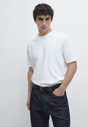 Giovane con capelli ricci scuri che indossa una maglietta bianca a maniche corte infilata in jeans blu scuro con cintura nera.