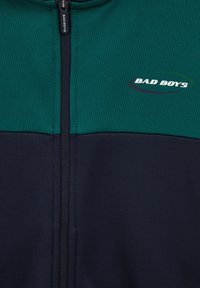 Jacka med dragkedja i grön och marinblå färgblockdesign. Har "BAD BOYS" logotyp i vitt och svart, tillverkad av texturerat tyg.