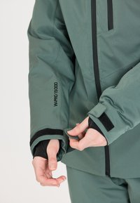 Grüne wasserdichte Jacke mit schwarzen Akzenten, verstellbaren Bündchen und sichtbarer Stoffstruktur. Auf dem Ärmel befindet sich ein Etikett mit der Aufschrift "WPRO 15000".