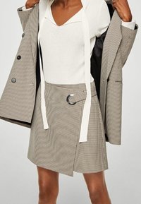 Falda marrón de cuadros con diseño envolvente, que cuenta con una hebilla metálica circular y un lazo blanco. Combinada con una blusa crema y un blazer gris.