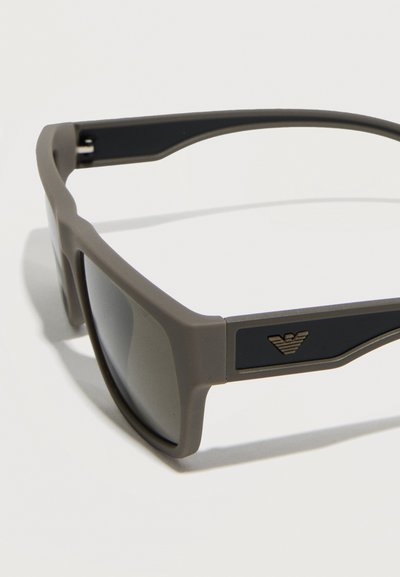 Ορθογώνια γυαλιά ηλίου σε ματ γκρι χρώμα με σκούρους φακούς και μαύρα πλαϊνά μπράτσα, που φέρουν ένα μικρό χρυσό λογότυπο αετού στη πλευρά.