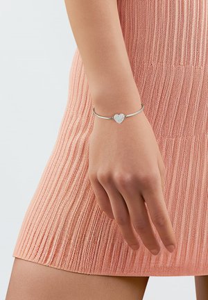 Zilveren armband met een hartvormige bedel, met een gestructureerd oppervlak, gedragen aan een hand. De arm van de drager is gedeeltelijk zichtbaar, naast een geribd roze kledingstuk.