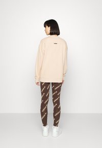 Gai beigts pārmērīgi lielais džemperis ar apaļu kakla izgriezumu, apvienots ar brūnām legingām, kurām ir atkārtota zelta logotipa raksts, un baltām kedām.