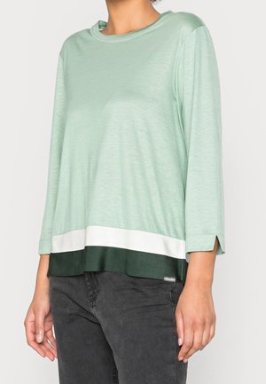 Persoon draagt een lichtgroen shirt met lange mouwen en een zoom in wit en donker groen kleurblok, gecombineerd met donkergrijze broek.