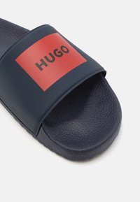 Mørkeblå slider sandal laget av teksturert gummi. Har en bred stropp med en rød rektangulær logo som viser "HUGO." Flatt, åpent tådesign.