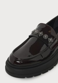 Mocassin brun foncé brillant avec semelle noire épaisse et clous argentés décoratifs sur la bride à l'avant.