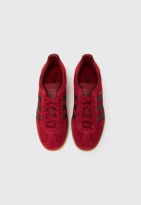 Piros velúr tornacipő fekete csíkokkal, texturált nyelvvel és gumitalpú gumitalppal. Megerősített orral és párnázott gallérral a kényelem érdekében.