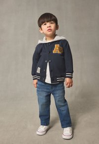Giacca varsity navy con cappuccio grigio, toppa ricamata a forma di "A", polsini a righe, abbinata a jeans blu e sneakers bianche. Dettagli in tessuto testurizzato.