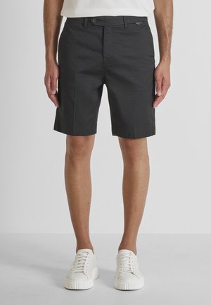 Hombre con pantalones cortos negros hasta la rodilla y zapatillas blancas, de pie contra un fondo gris claro liso.