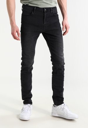 Homme portant un jean noir slim et des baskets blanches, debout devant un fond blanc uni.