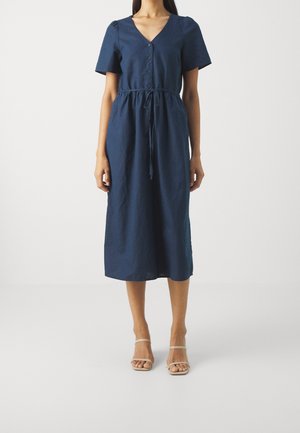 Kvinde iført en navyblå midi-kjole med knapper, korte ærmer og bælte i taljen, kombineret med beige åbne hæle, stående på en ensfarvet baggrund.