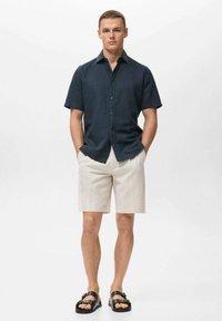 Camisa de manga corta color azul oscuro, pantalones cortos beiges y sandalias negras. La camisa presenta un diseño de botones y un ajuste relajado.