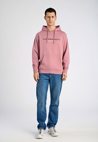 Sudadera rosa con un bolsillo frontal y cordones, que presenta un logo impreso de "Copenhague", combinada con unos jeans azules y zapatillas blancas.