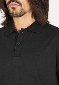 Svart polo skjorta gjord av texturerat tyg, med krage, tvåknappars placket och en subtil präglad logotyp på bröstet.