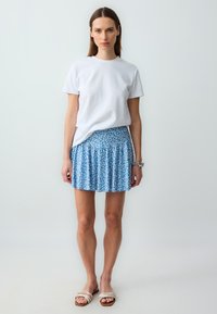 Weißes T-Shirt mit Rundhalsausschnitt kombiniert mit einem blauen Minirock mit Blumenmuster, der Falten und einen strukturierten Stoff aufweist. Getragen mit beigen, schnürlosen Flatsandaletten.